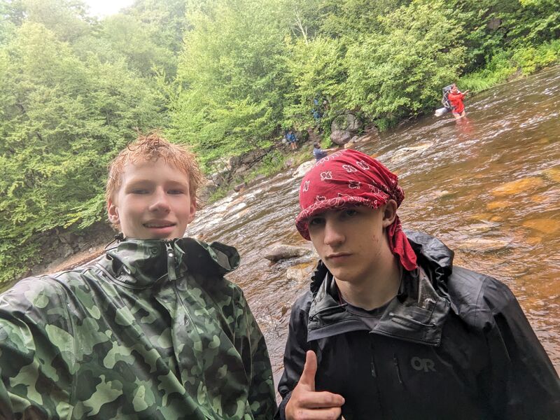 Two young men stand close to the camera, seemingly posing for a picture. The person on the left has red hair and is wearing a camouflage jacket. The person on the right is wearing a black jacket and a red bandana. They appear to be outdoors, possibly near a rocky, wet area with a stream or waterfall in the background. Other people can be seen further up the slope in the background.